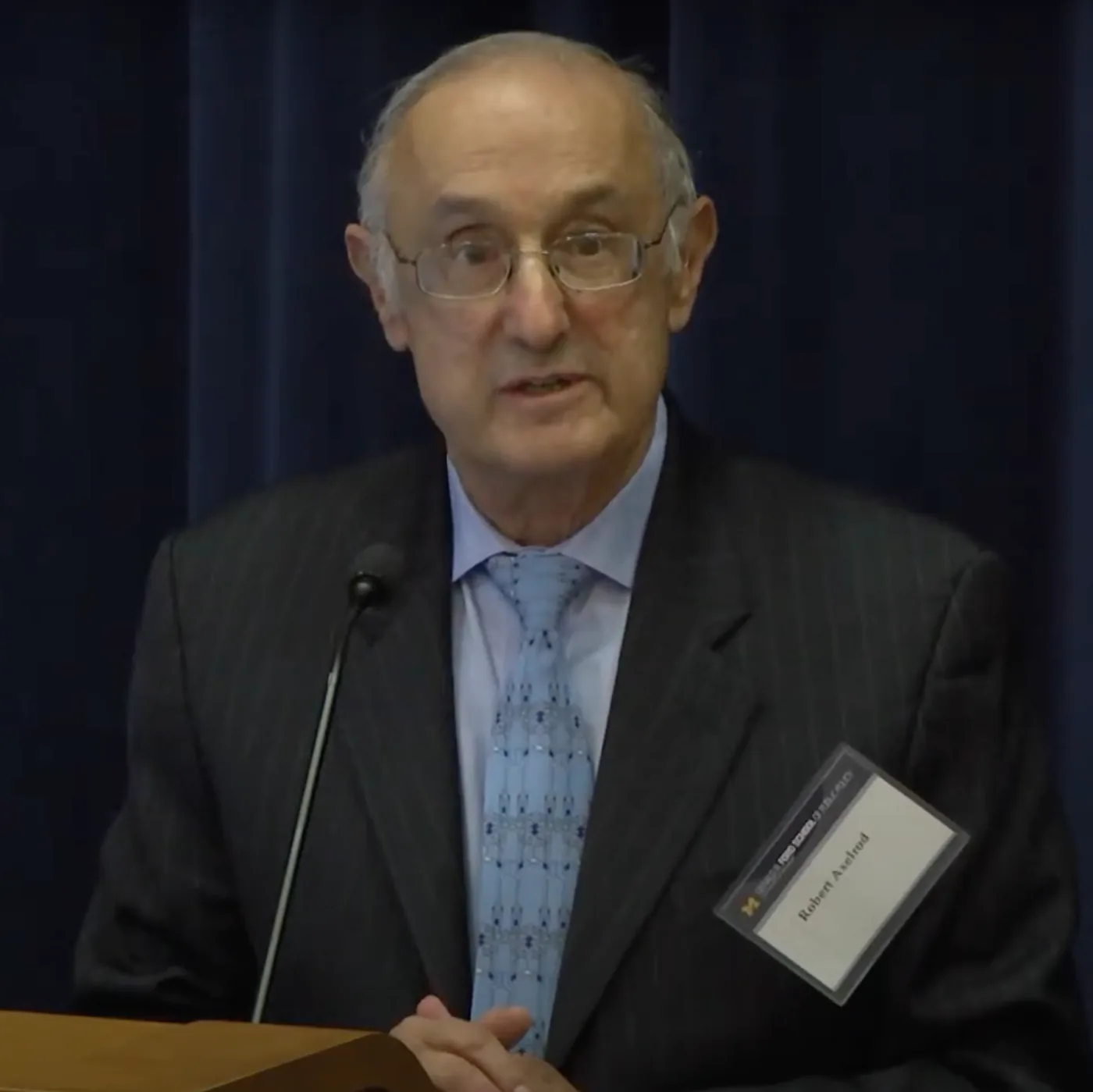 An elderly man in glasses and a dark suit speaking behind a lectern at a microphone