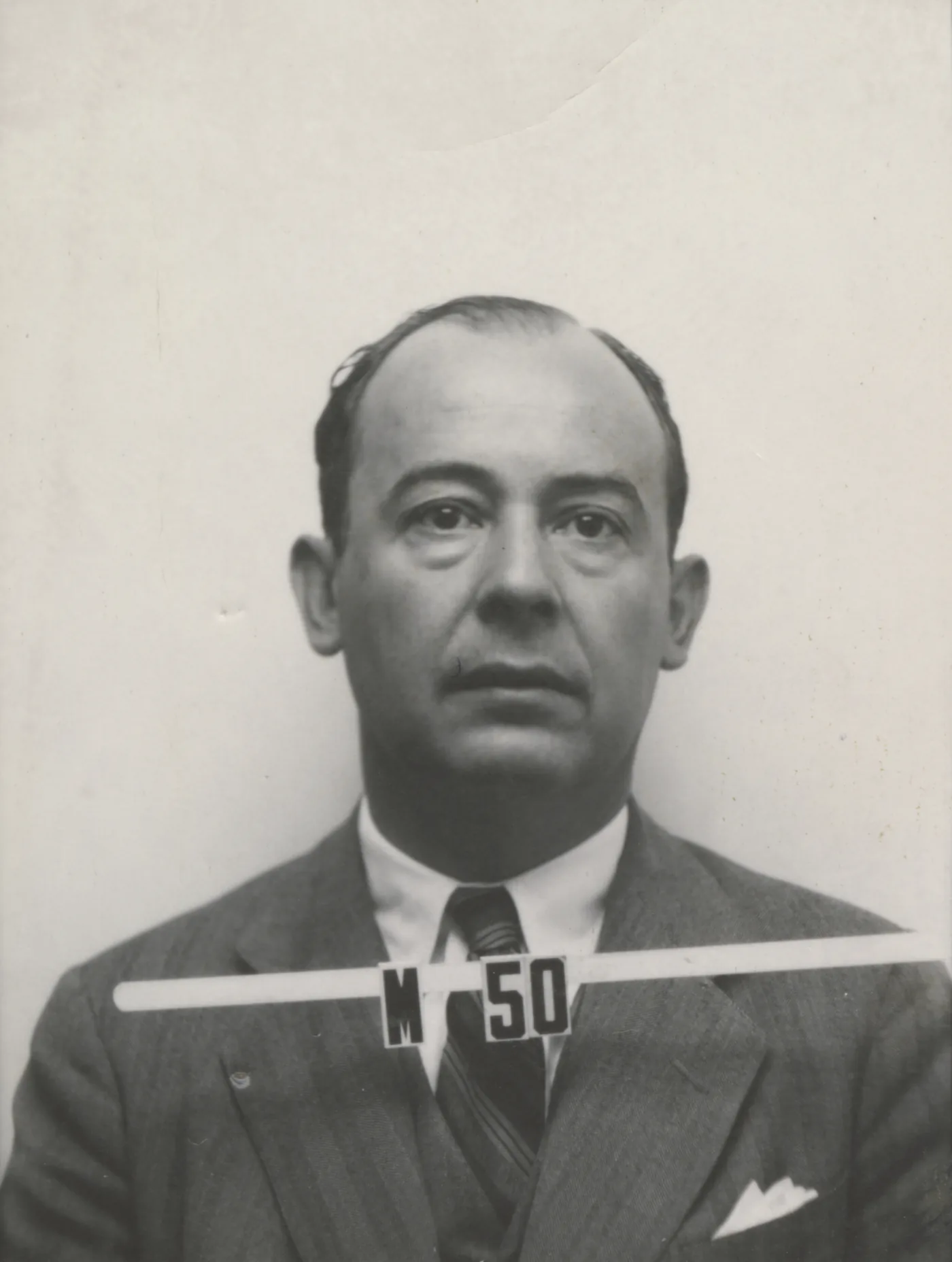A black-and-white identity badge photograph of a man in a suit and tie, looking at the camera